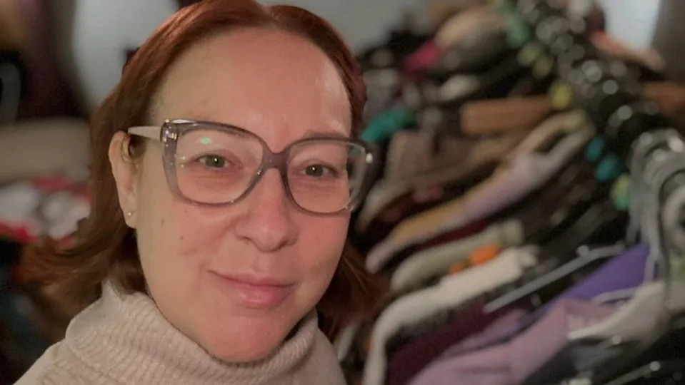 Selfie photograph of Rachel Grady. She has a ginger short bob, wears square glasses, has blue eyes and smiles at the camera. The background is blurred but shows railings of clothes.