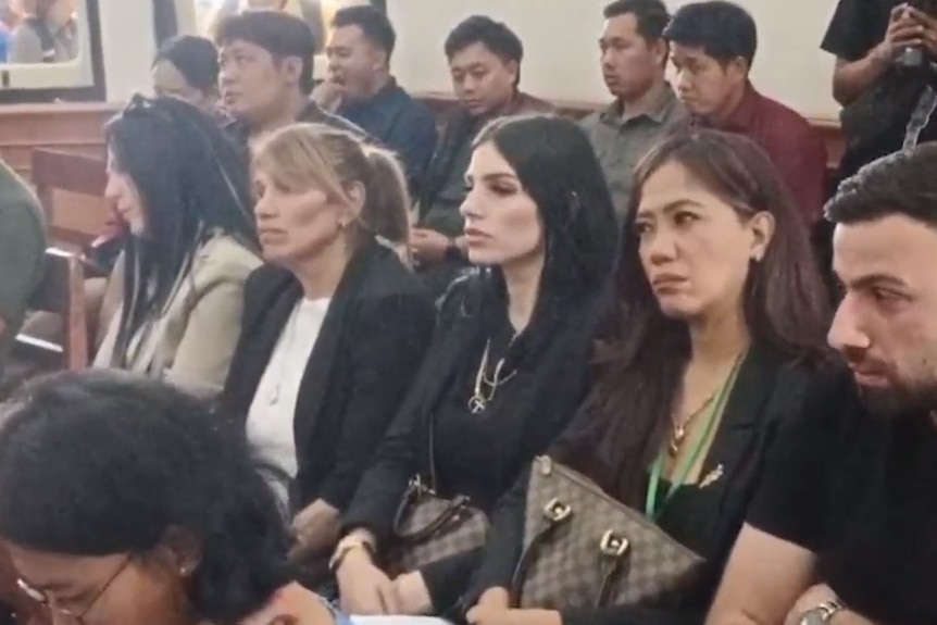 A young woman with long black hair sits in a public gallery inside a court room.