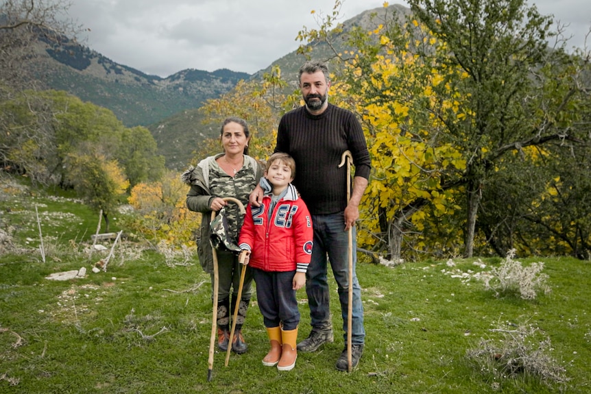 A family of three, each with a shepherd's crook, standing in front of mountains. 