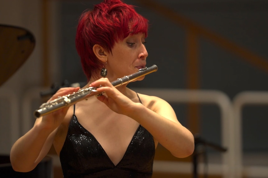 Eliza Shephard plays a silver flute in a wooden concert hall. She has short red hair and wears a sparkly black top.