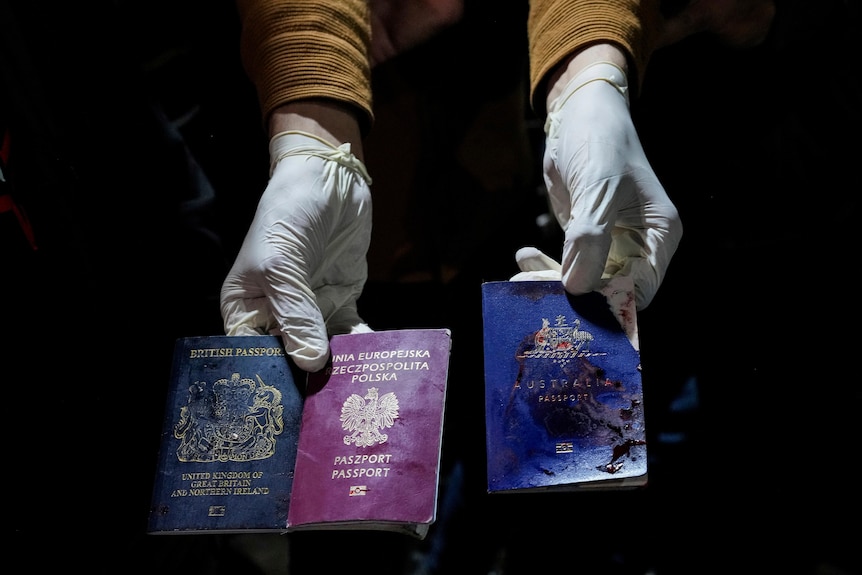 Gloved hands holding up three bloodied passports. One is British, one Polish and one Australian.