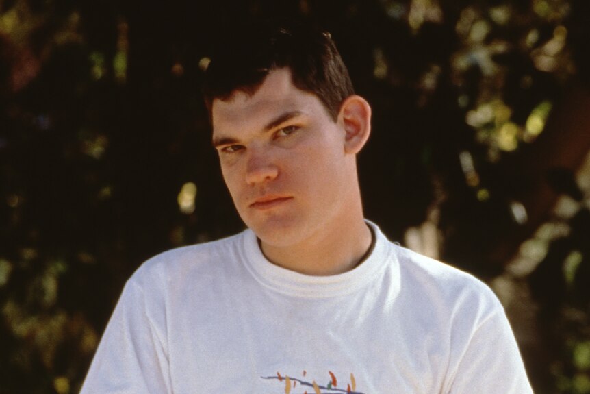 A teenager stands in a white T-shirt and jeans holding a BMX bike. A yellow 'L' plate is on the front and the boy is standing
