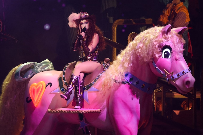 Wearing a cowboy hat and sparkly outfit, Chappell Roan sits on a large pink carousel horse and sings into a microphone.