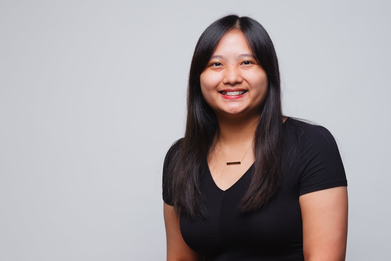 Head and shoulders portrait of young woman with long, straight black hair. She has braces on her teeth and is wearing a V-neck T-shirt. 