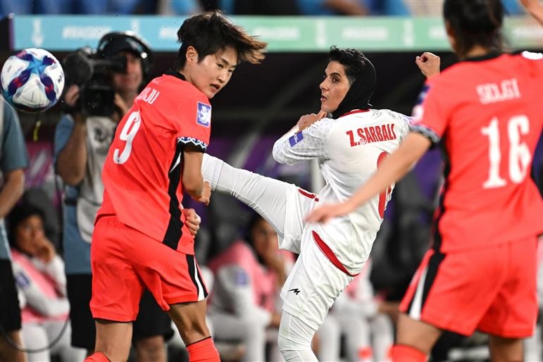A Middle Eastern woman in white jersey and tudung kicks past red-shirted Asian women in soccer game 