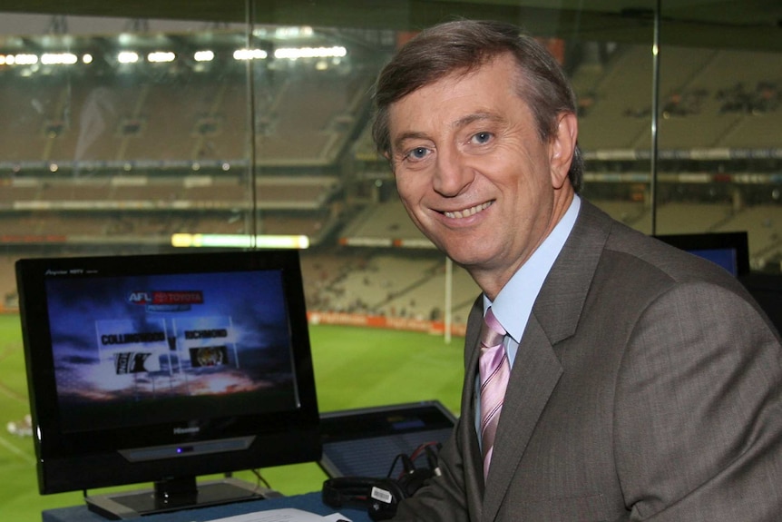 A smiling, middle-aged man in a suit sits in a commentary box overlooking a sports field in a stadium.