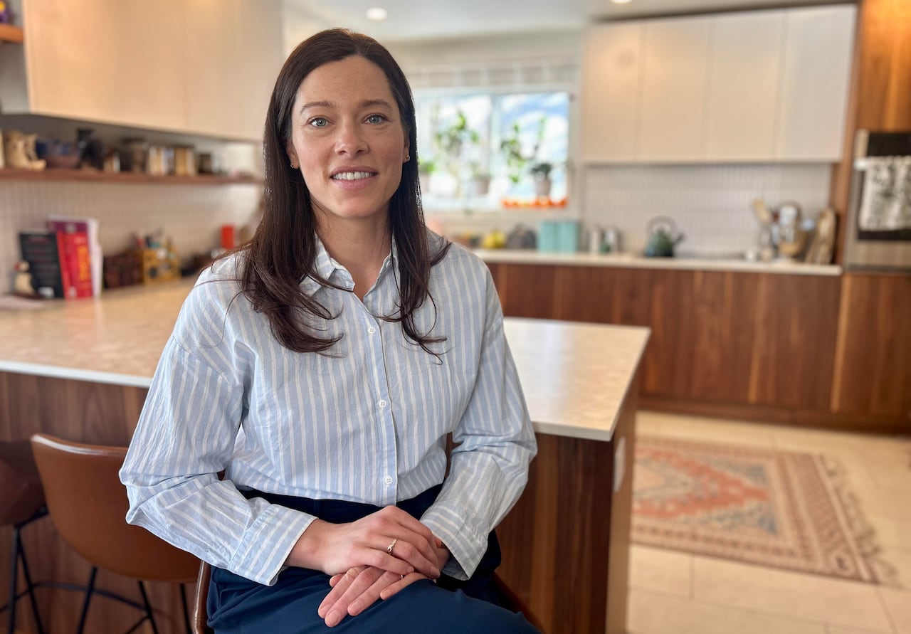 A woman with long brown hair smiles at the camera while sitting on a stool in a kitchen