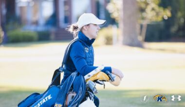 Alt text:  Veronika Kedronová of Kent State women’s golf poses with the Lady Luck Invitational individual champion trophy alongside a UNLV staff member in a red quarter-zip and white UNLV hat. Kedronová wears a white Kent State polo, blue pants and a navy cap as both smile and hold the large poker-chip-shaped trophy outdoors in Las Vegas.