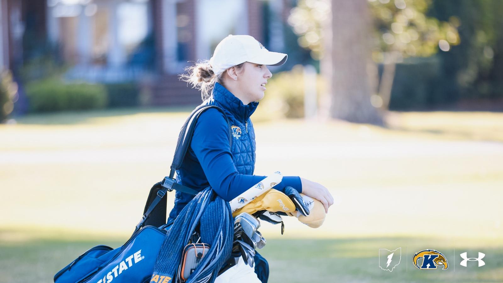 Alt text:  Veronika Kedronová of Kent State women’s golf poses with the Lady Luck Invitational individual champion trophy alongside a UNLV staff member in a red quarter-zip and white UNLV hat. Kedronová wears a white Kent State polo, blue pants and a navy cap as both smile and hold the large poker-chip-shaped trophy outdoors in Las Vegas.