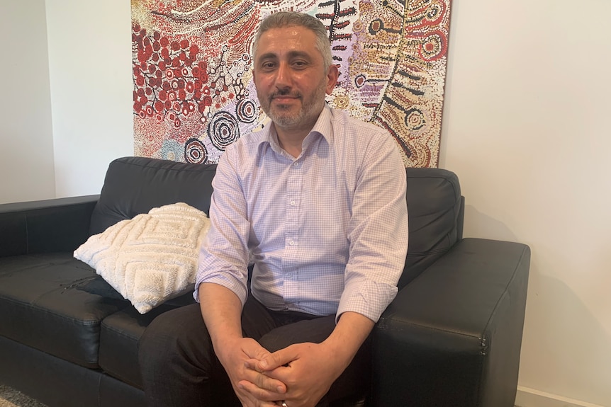 A man sitting on a sofa in front of an Aboriginal dot painting