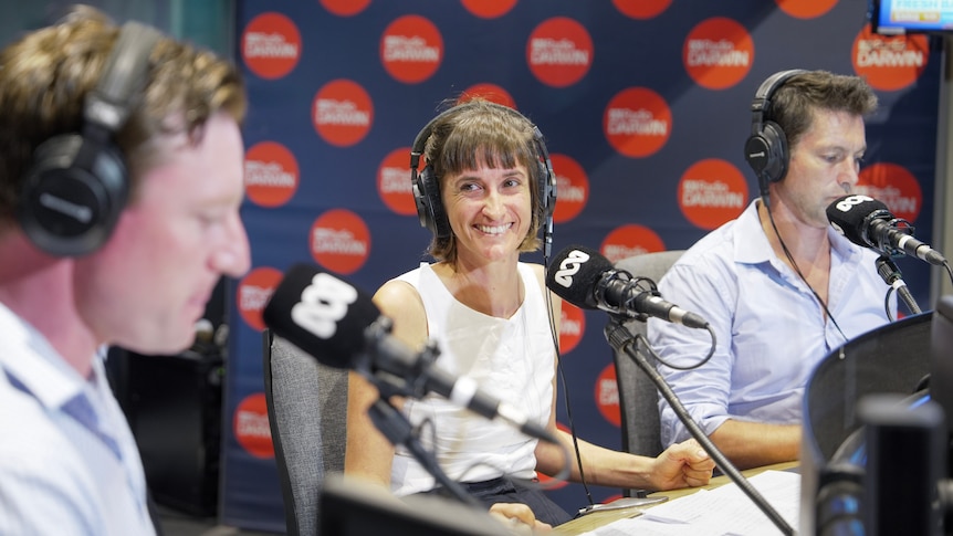 A woman, smiling widely, sitting in front of a microphone inside a radio studio, flanked by two men.