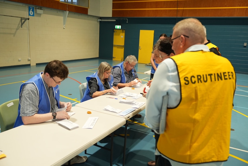 Counters sit at tables counting piles of ballots while scrutineers in yellow vests watch on