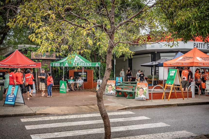 Three tents at the markets, including a red Labor tent, a Greens tent and an orange CLP tent. Trees are in foreground.