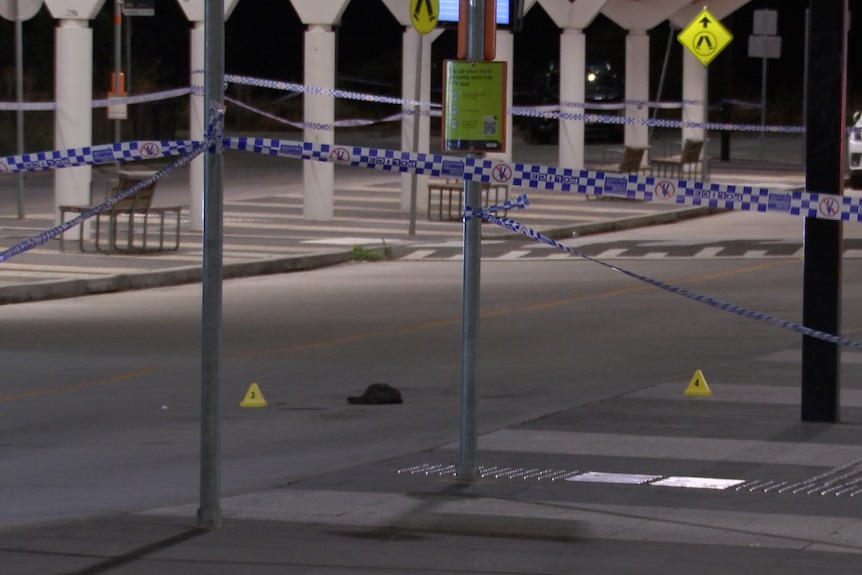 A dark cap lies between two yellow triangles on a concrete roadway near a pedestrian crossing, police car and police tape.