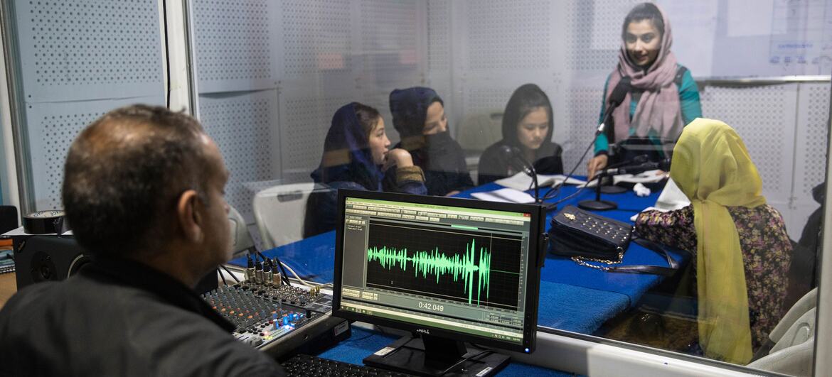 A female radio host and several young girls in a radio studio in Afghanistan, being monitored by a male audio engineer at a computer showing a sound wave.
