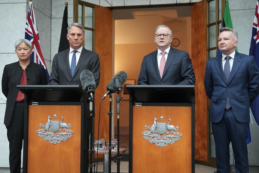 Two men and a woman in suits stand at a podium speaking to journalists with the Australian flag in the background.