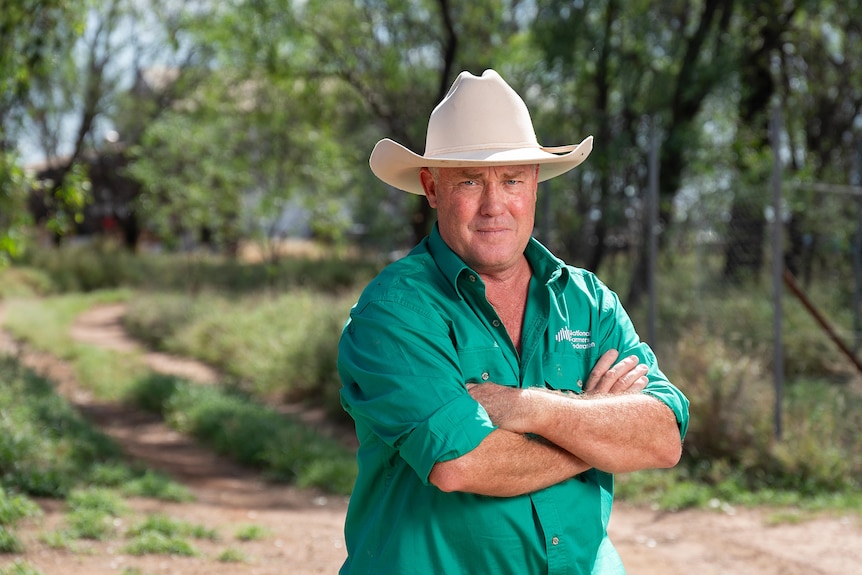 A man in a green shirt and broad white hat, standing outdoors with his arms crossed.
