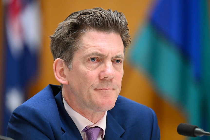 A midddle-aged man with dark hair wears a suit as he sits behind a microphone.