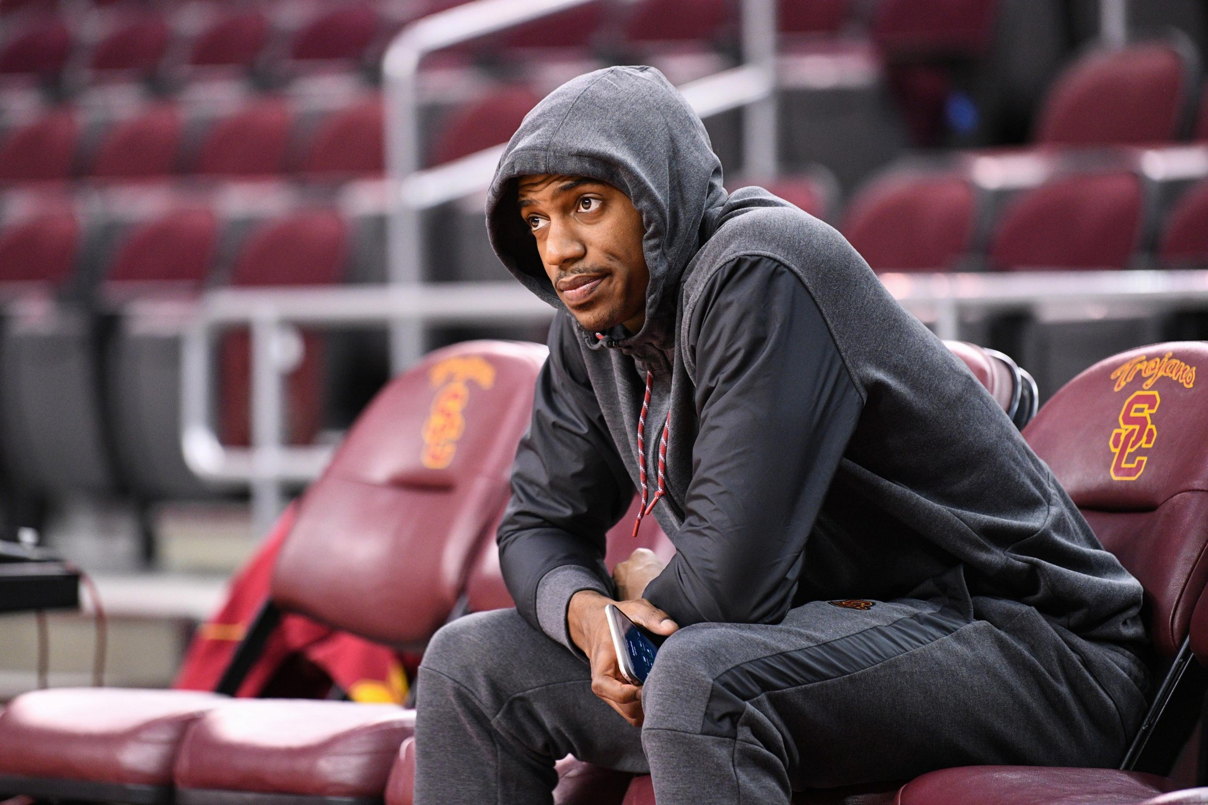 A man in a gray hooded sweatshirt and sweatpants sits on a USC bench, holding a phone and looking thoughtfully to the side.