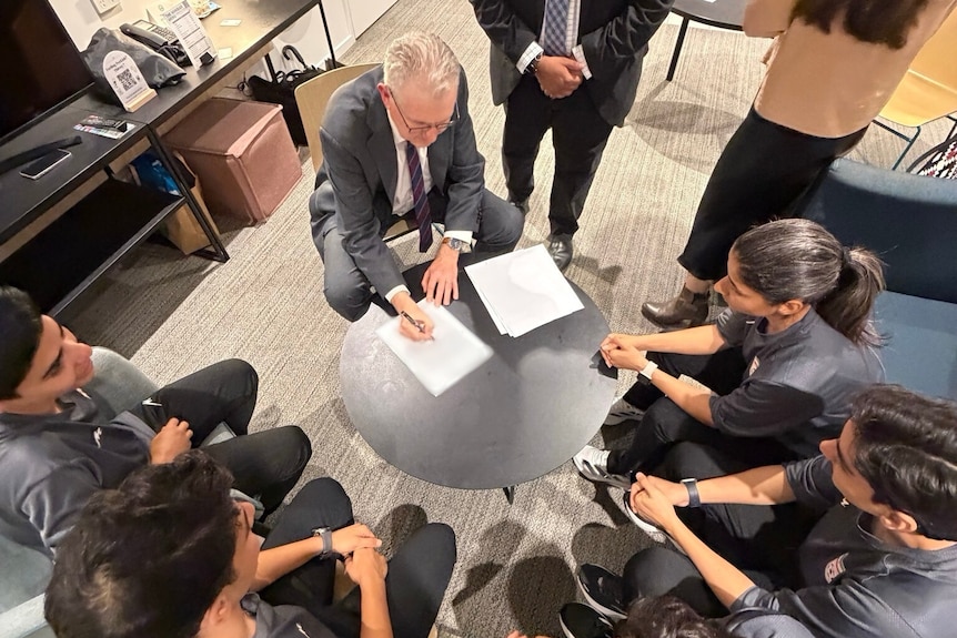 Tony Burke signing papers at a table, surrounded by the Iranian football team.