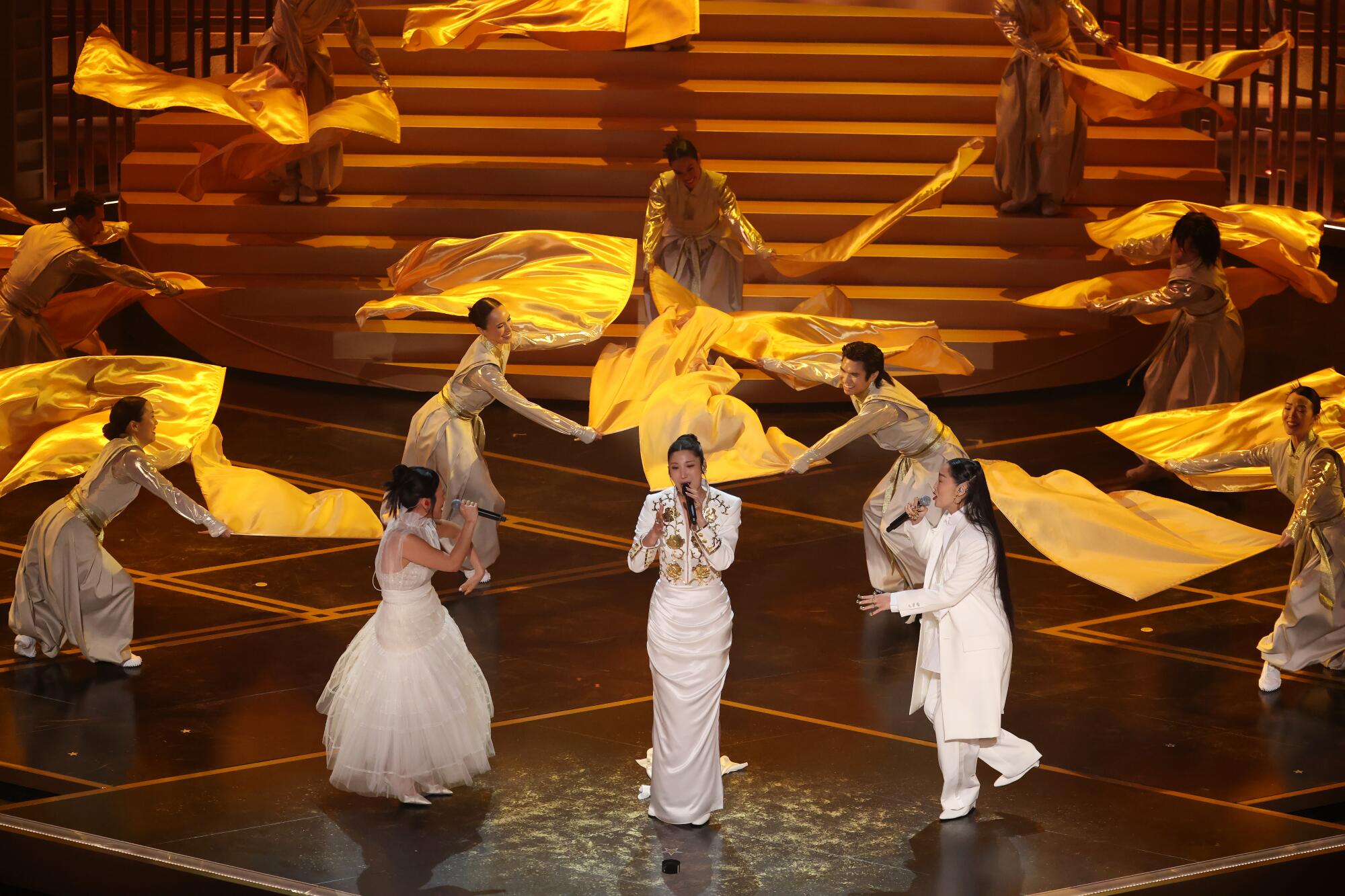 Three women in white and gold singing on stage with dancers around them.