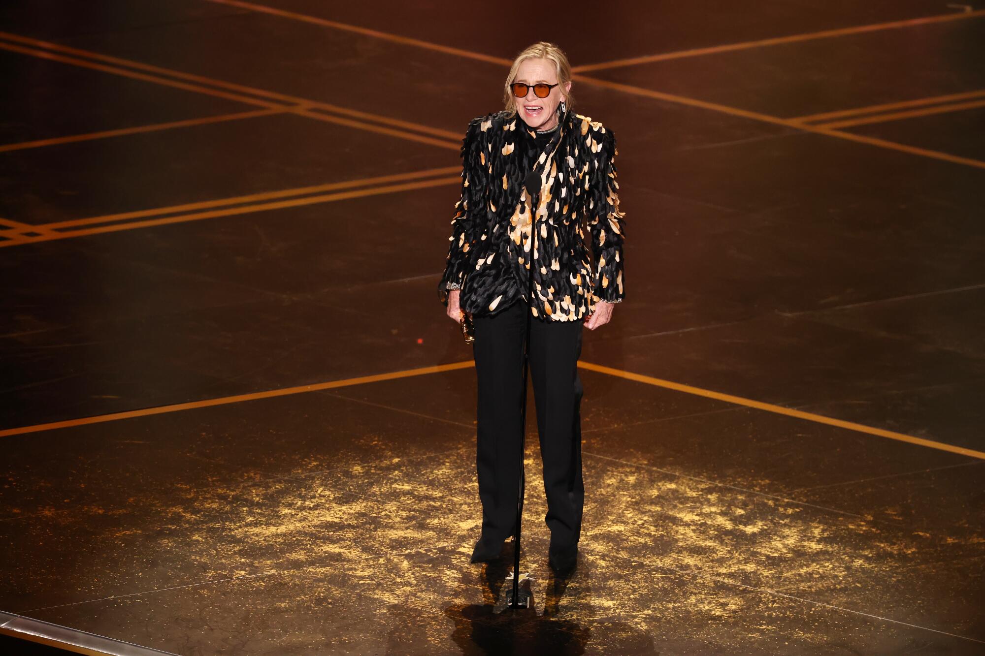 A woman in glasses, a black and golden blazer and black slacks speaks into a microphone on stage.