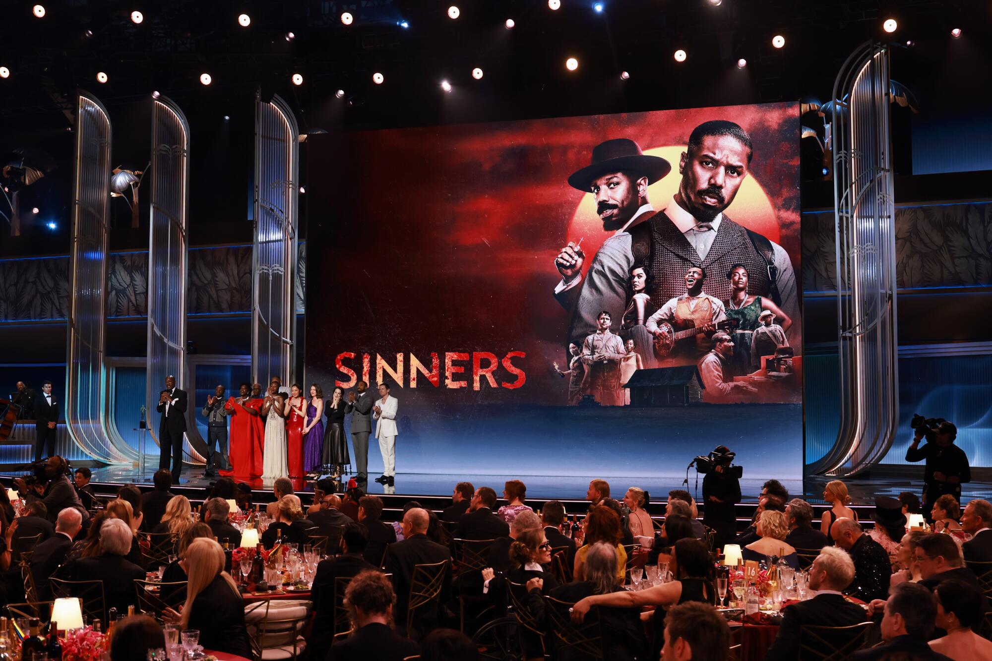 A group of people in formal wear standing onstage before a screen with an image of several people and the word "Sinners."