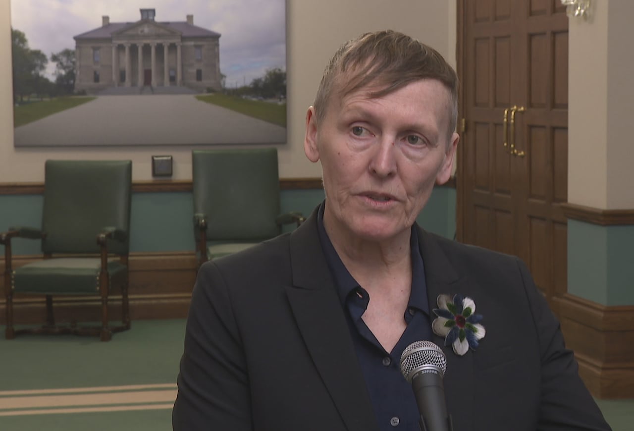 A woman with short hair, wearing a black suit, stands in front of a microphone in provincial government room.
