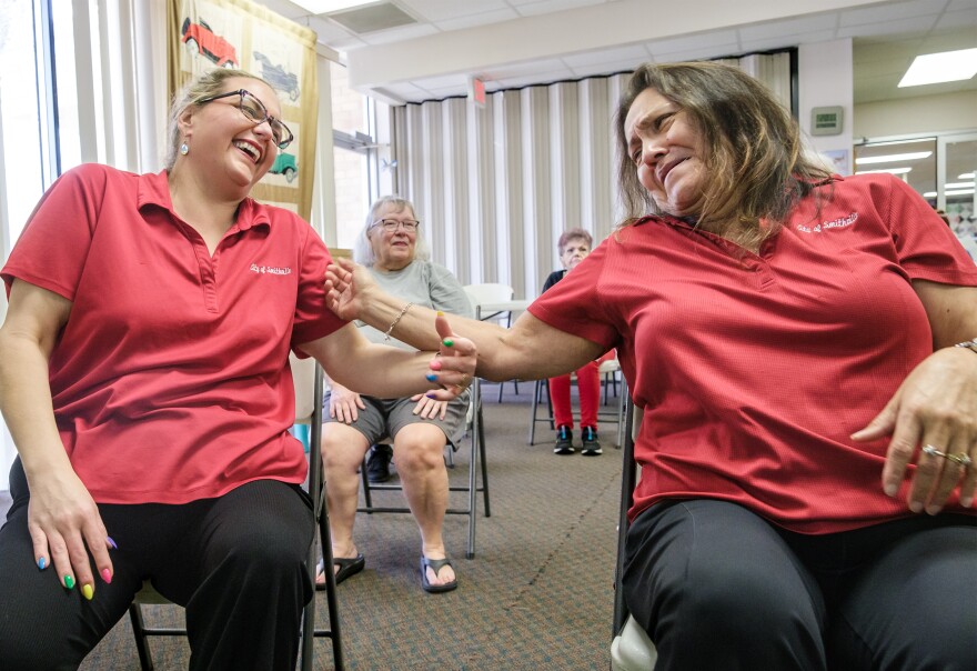 Two women wearing red shirts sitting next to each other reach out an arm toward the other one as they laugh. 