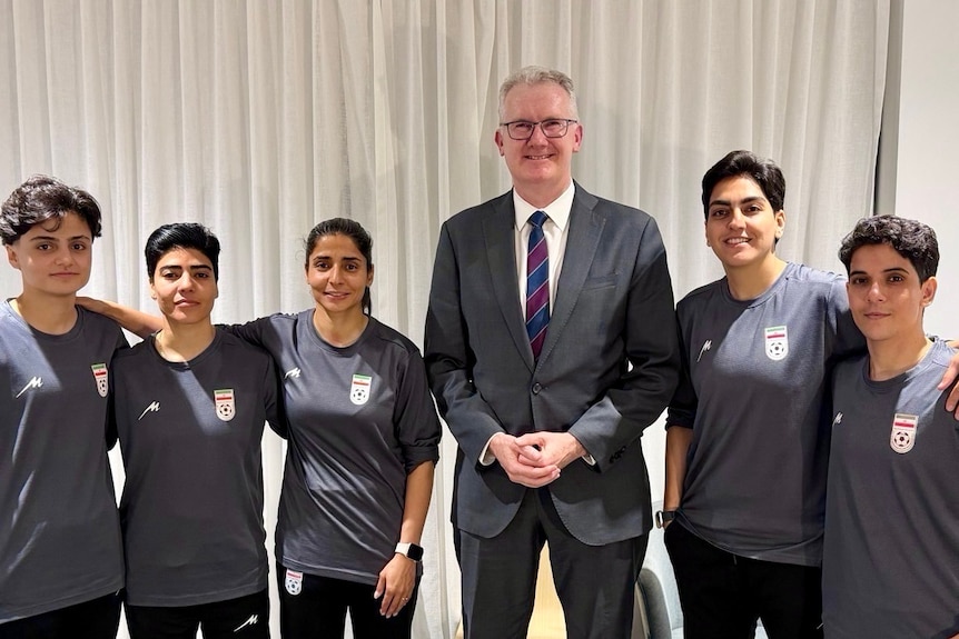 Mr Burke, a tall white man, towers over five Iranian women in t-shirts with matching team logos. 