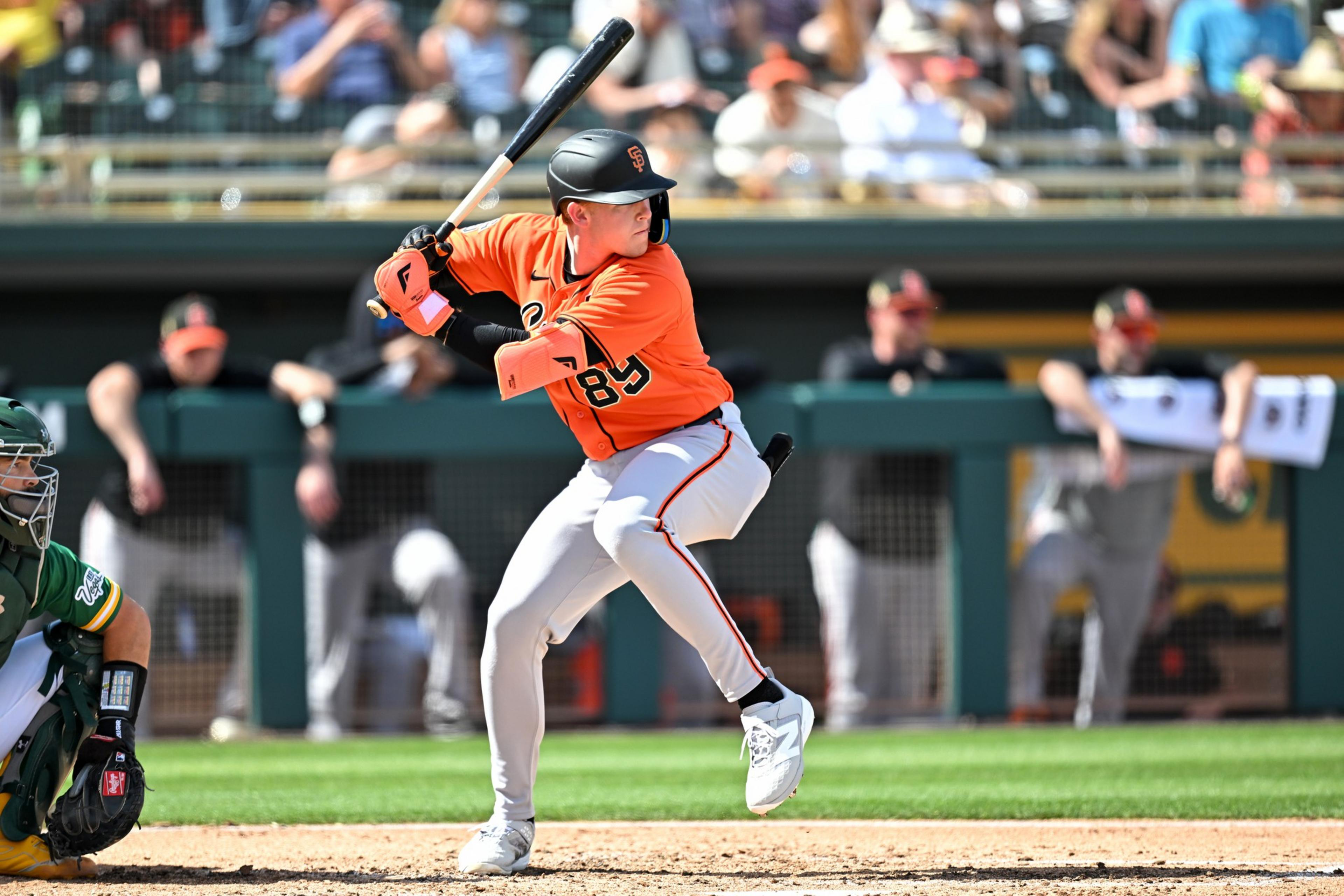 A baseball player in an orange jersey and gray pants is poised to swing at a pitch, with a catcher crouched behind him and spectators in the background.
