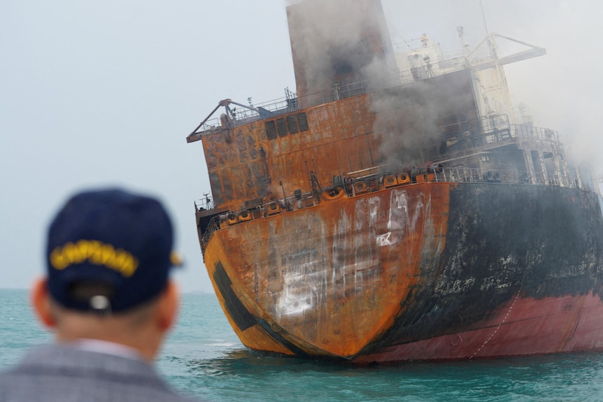 A man looks at a large burnt ship in the water, with smoke drifting out.
