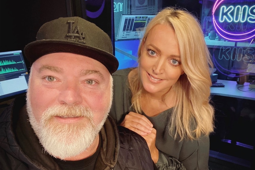 A man with a grey beard and black cap and a blonde woman resting her hand on his shoulder smile in a radio studio.