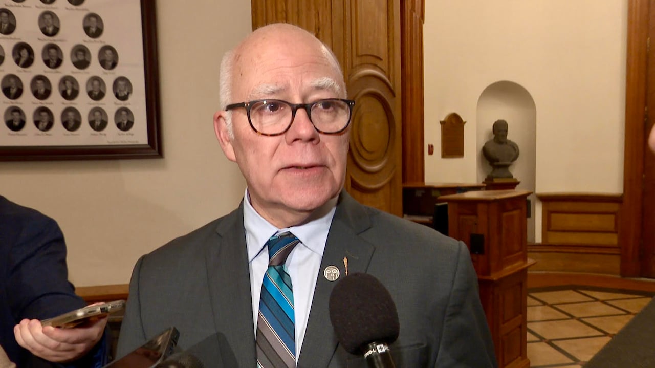 A man with white hair and a grey suit looks to his left with microphones pointed at his mouth.