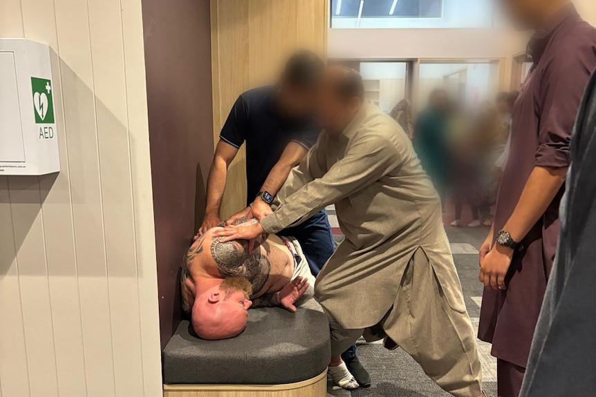 Two men forcibly restrain a topless tattooed white man.