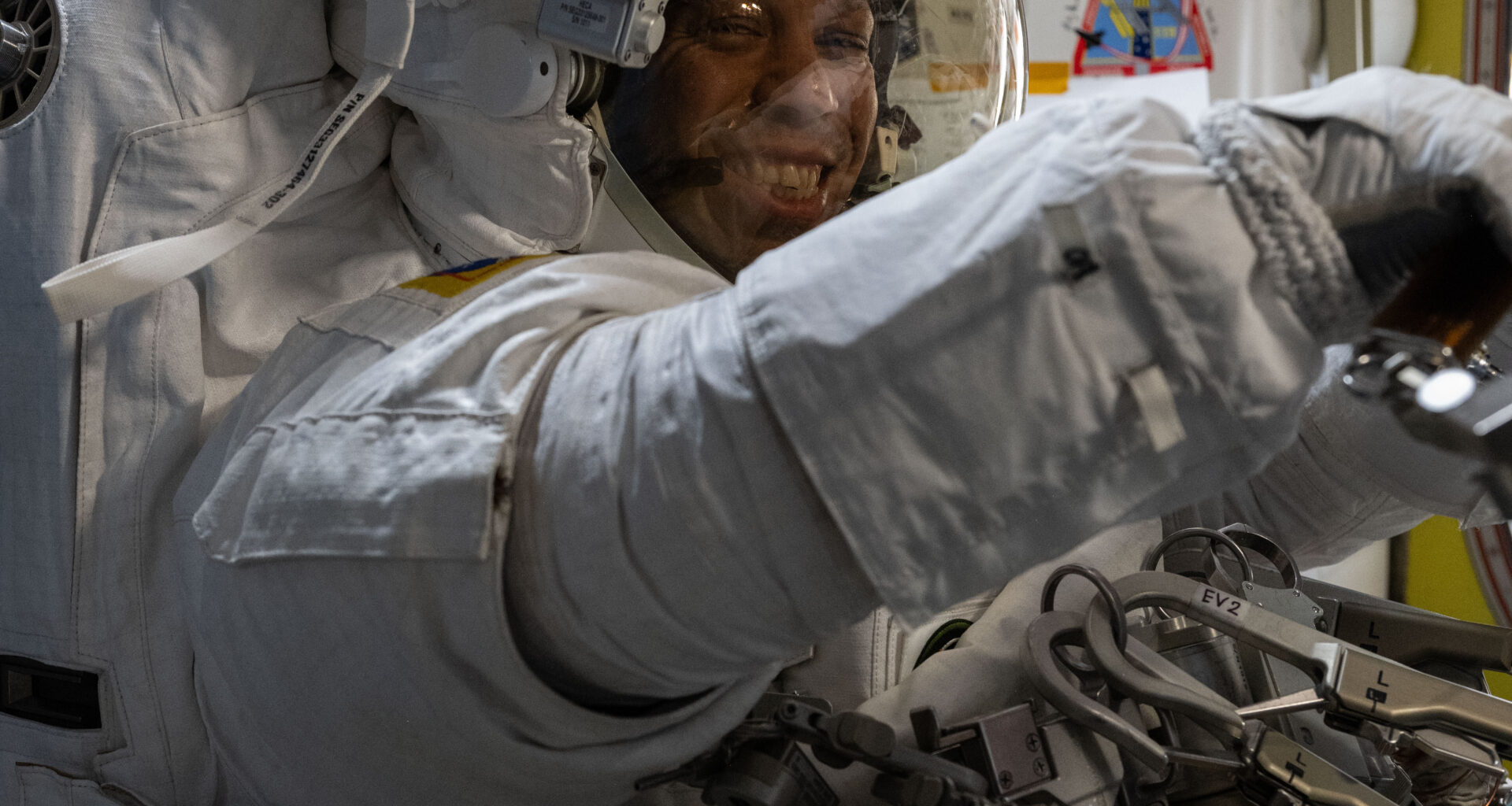 Chris Williams, an astronaut, is wearing a white spacesuit and helmet. He is facing the camera, but his body is turned to the right of the image. His arms are extended in front of him.