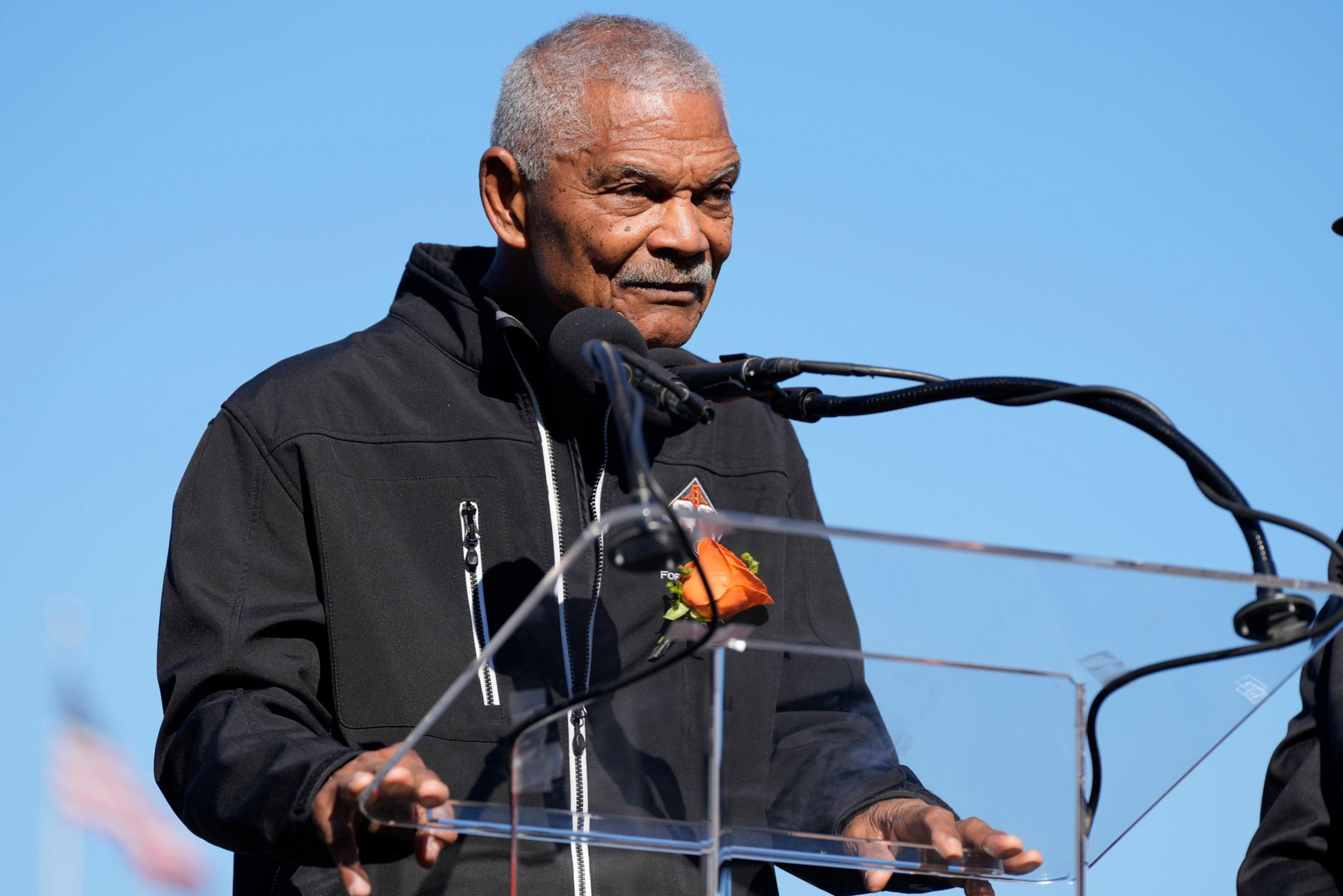 An older man with gray hair and a mustache speaks into microphones while standing behind a clear podium, wearing a black jacket adorned with an orange flower.