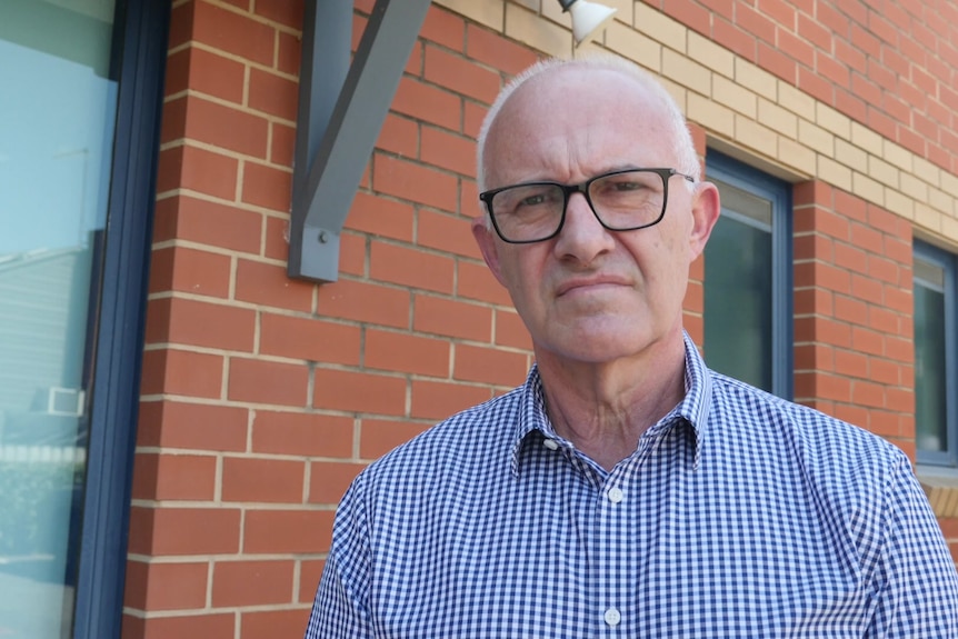 A man with a stern look on his face stands outside a red brick building.