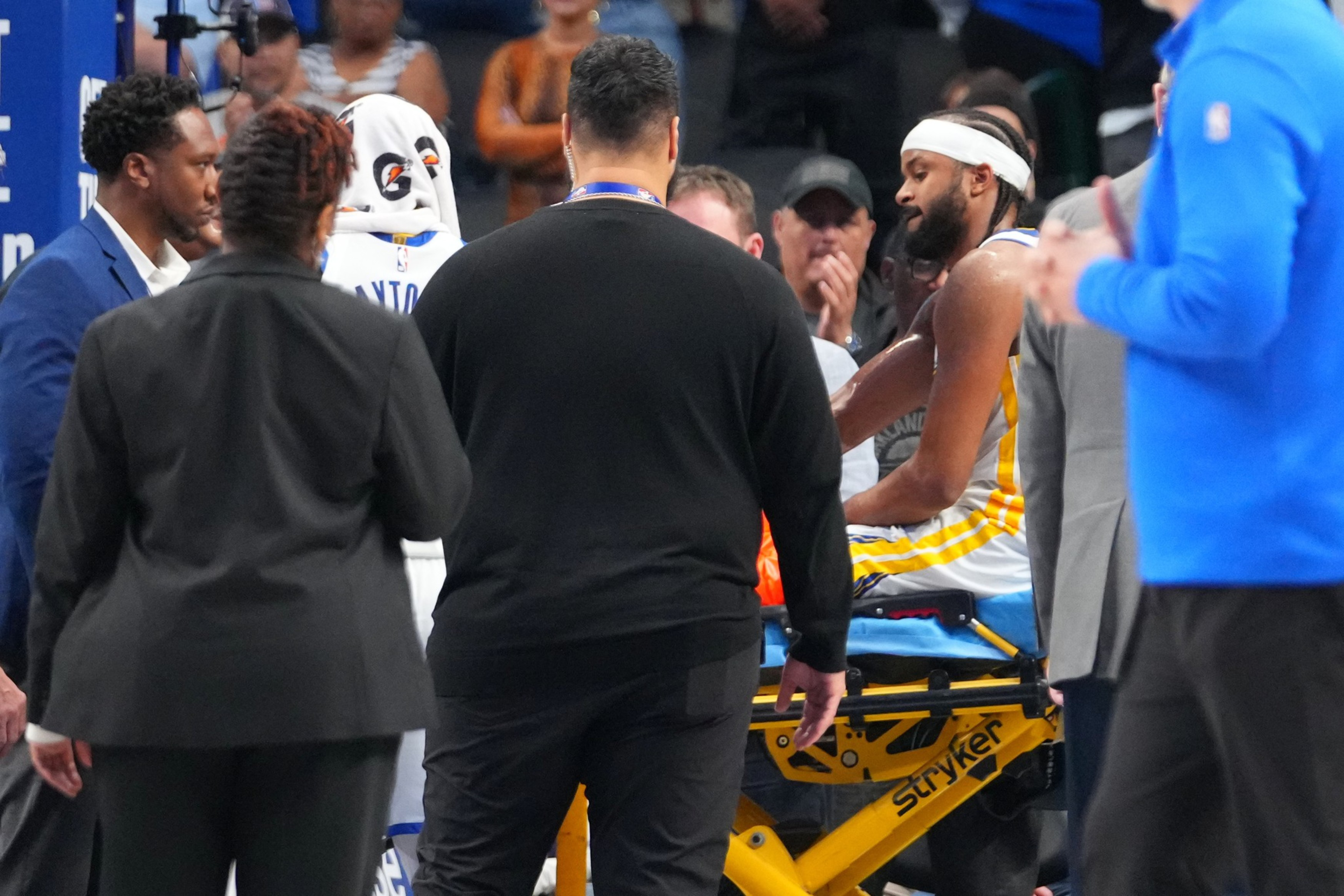 A basketball player in a yellow and white uniform with a headband sits on a stretcher, surrounded by people in suits and casual clothes.