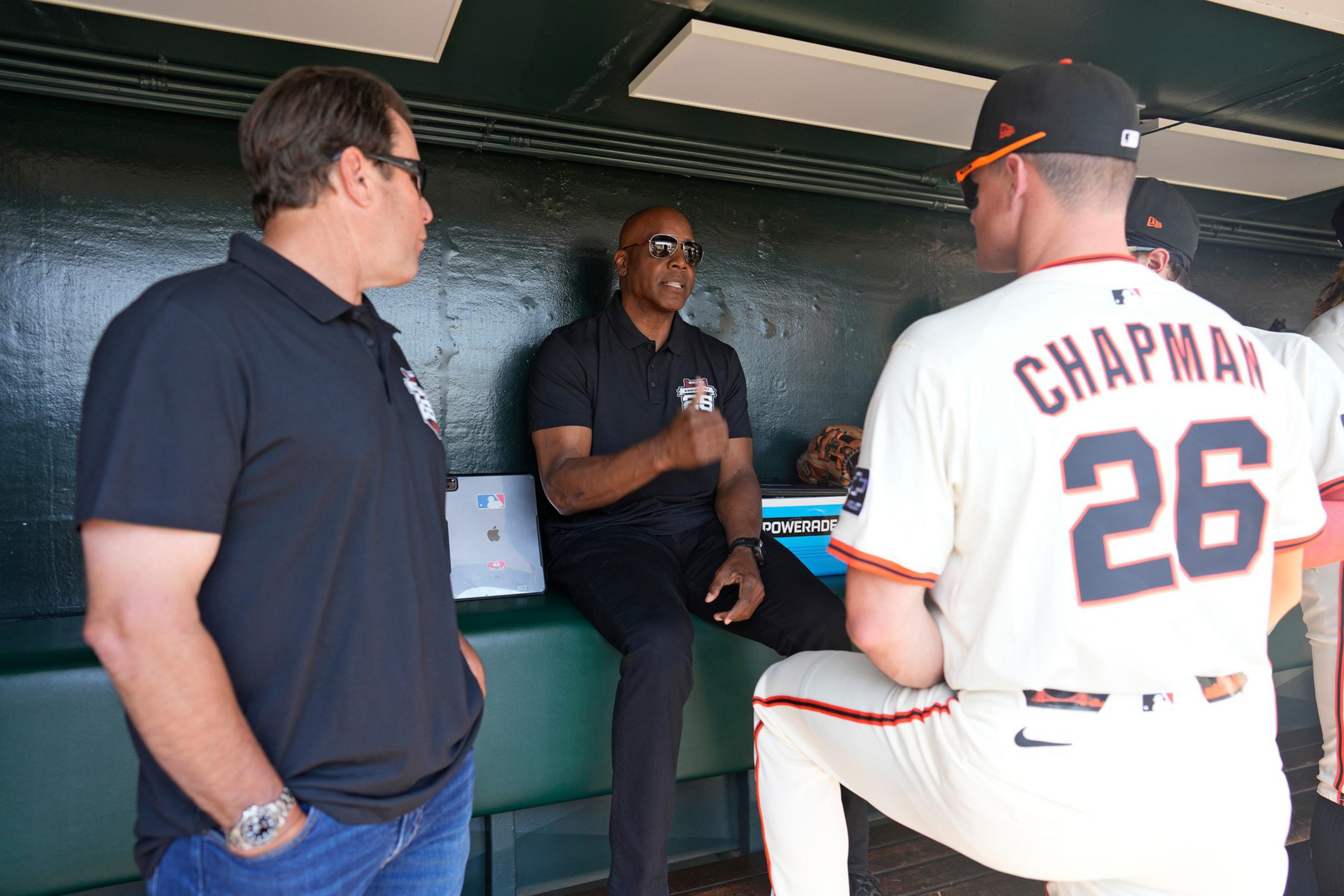 Three men in a baseball dugout are talking; one in a black polo and sunglasses is seated while two others, one wearing a black shirt and the other a white jersey, stand nearby.