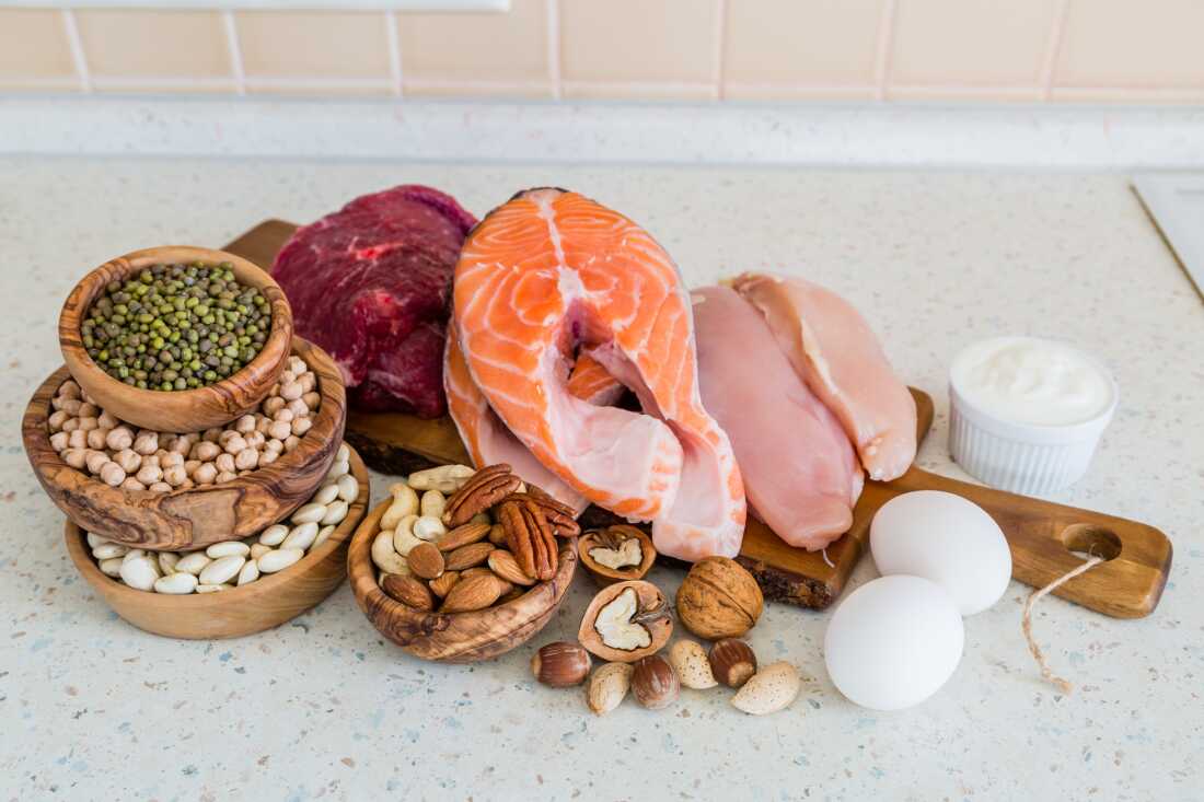 A selection of nuts, beans, eggs, raw chicken, fish and beef are displayed on a wooden board and in bowls sitting on a counter.