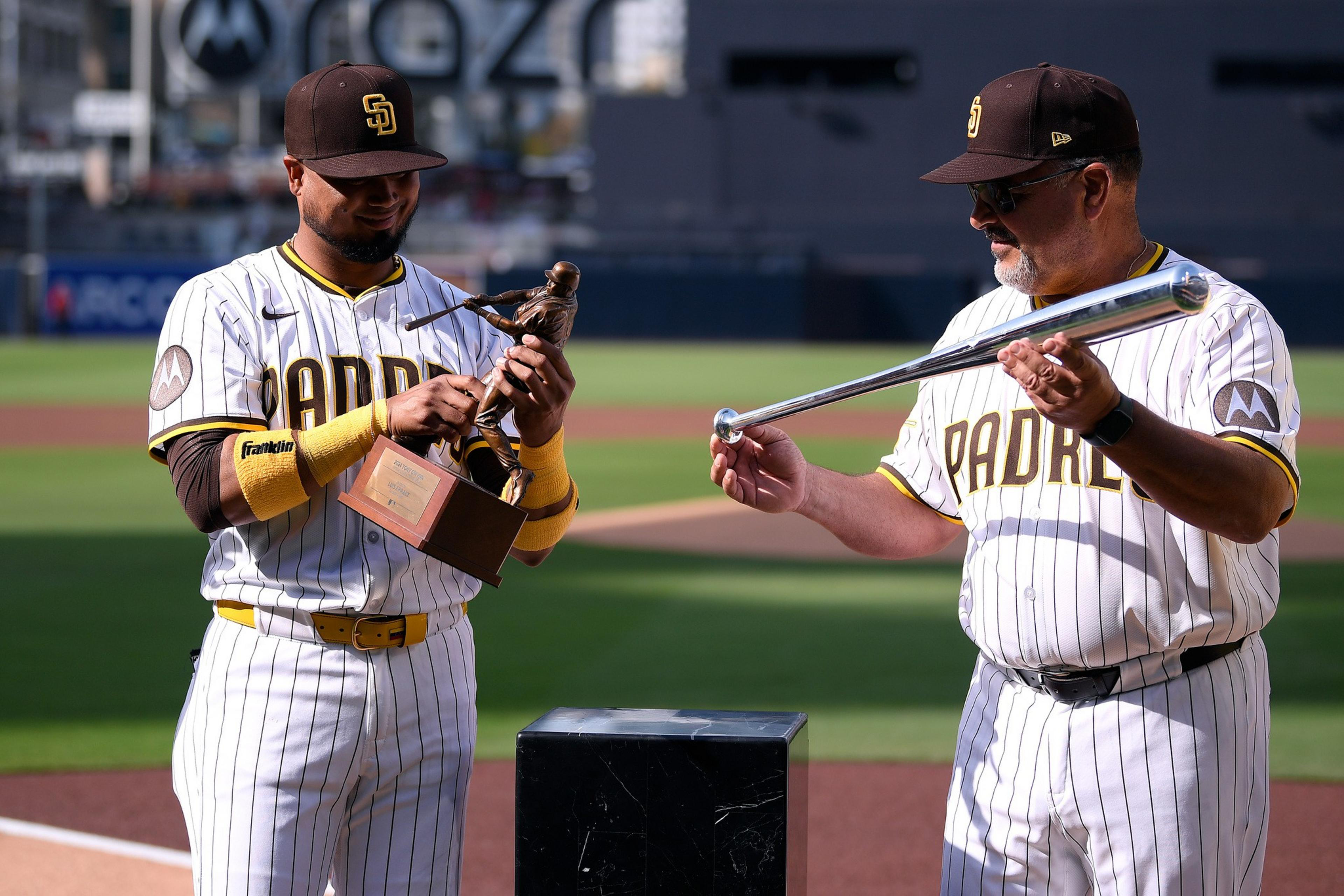 Two San Diego Padres baseball players in uniform hold a trophy and a bat on a baseball field during daylight.