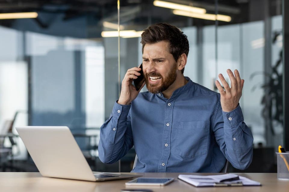 Angry and irritated young businessman sitting in the office at the desk and talking on the phone while waving his hands