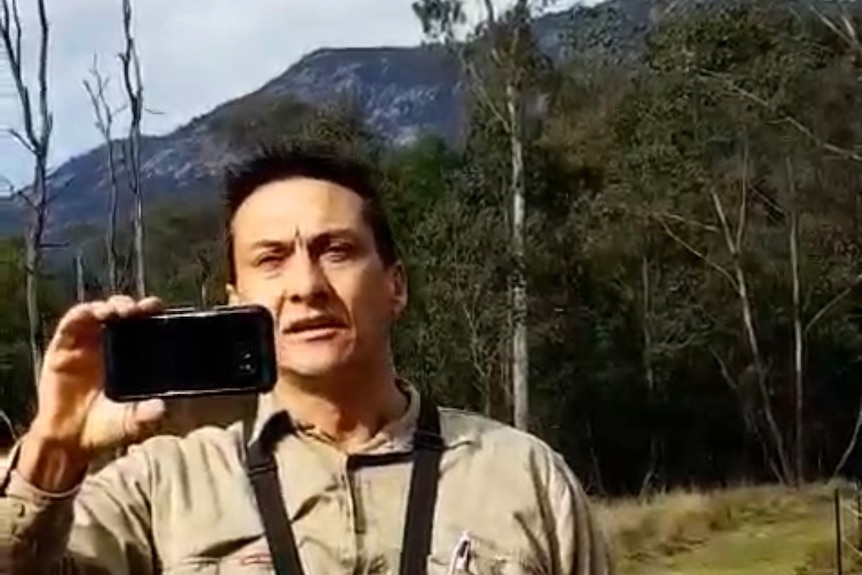 Man holding phone horizontally while wearing body camera on a property that looks like a farm with mountain in the background
