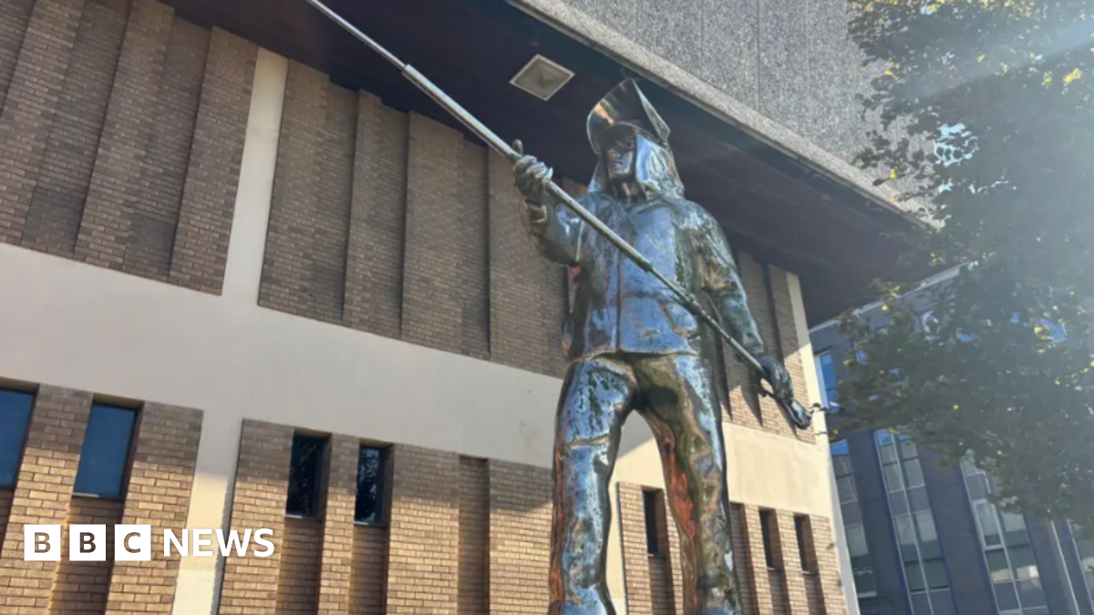 A metal statue showing a life-sized man dressed in steelworkers' uniform and holding a long pole with both hands. There is a light-coloured brick building in the background.