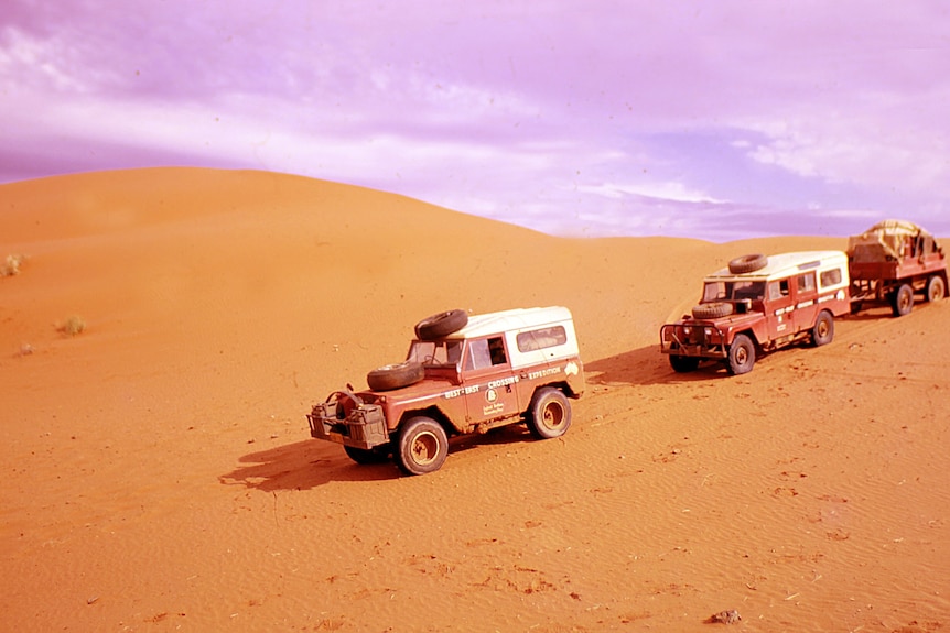 Three red four-wheel-drives travel in a line across hilly red dirt of the outback