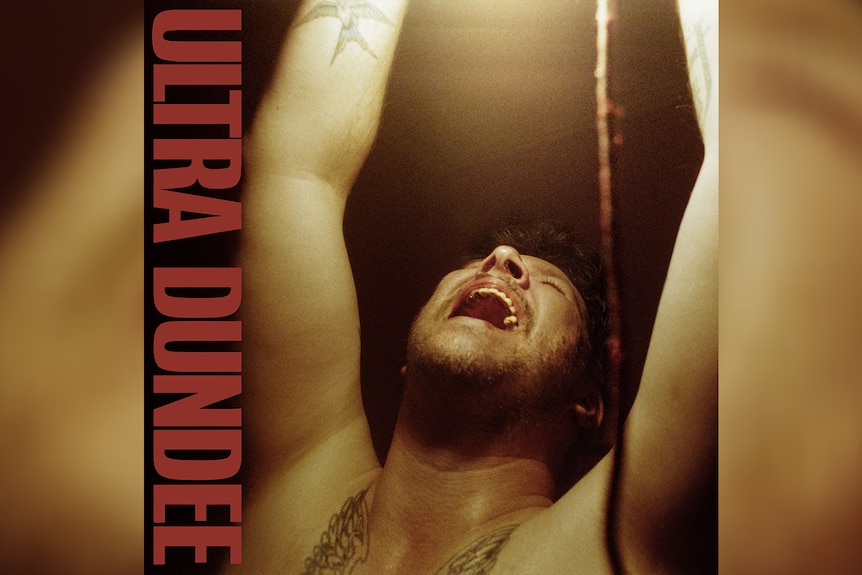 shirtless middle-aged white man with tattoos stands under light, arms up and shouting. Vertical text on left reads: ULTRA DUNDEE