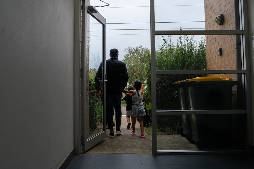 A man and his two young children can be seen from behind, walking out the glass doors to a building.