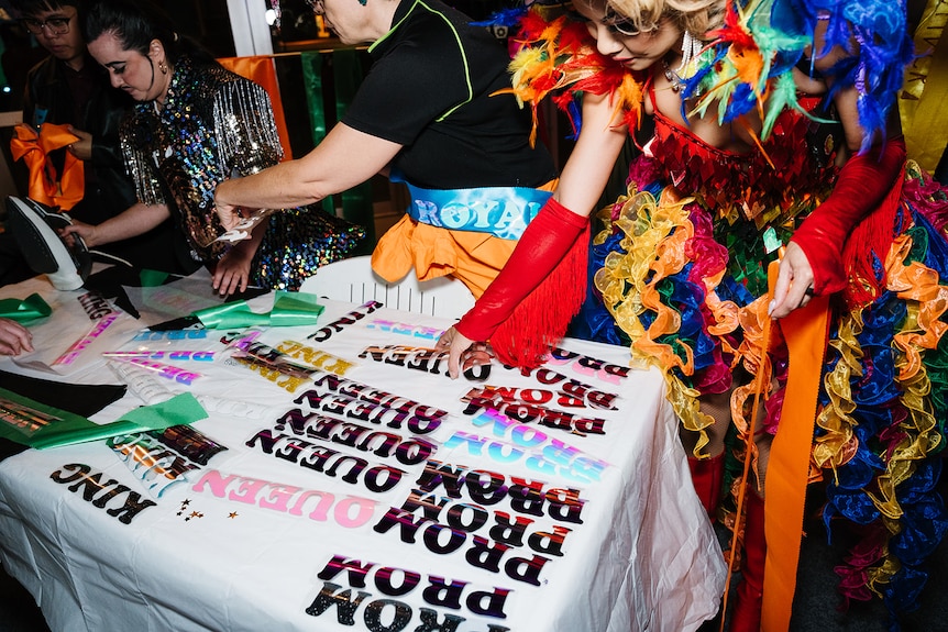 A person in rainbow chiffon reaches down to a table with the words 'prom'