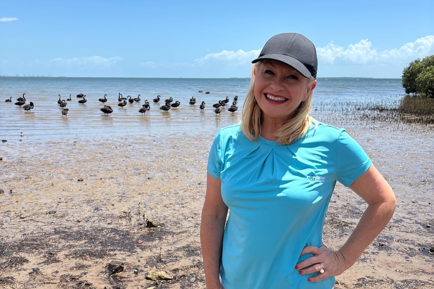 A blonde woman in a blue t-shirt standing on the beach with a flock of black swans behind her, she is smiling at the camera.
